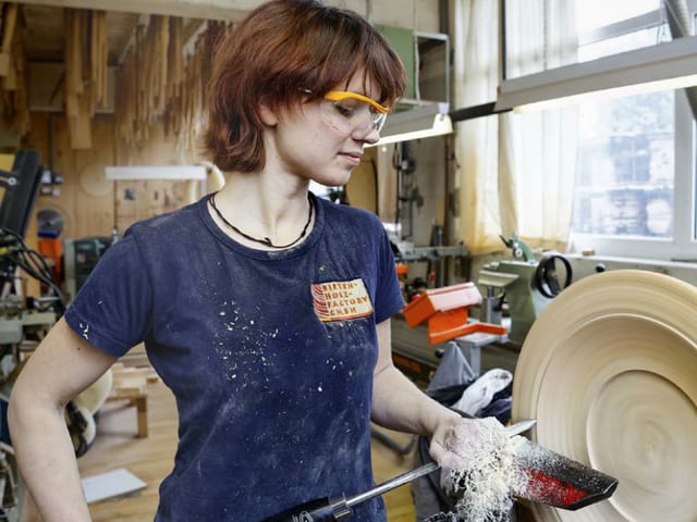 Die Holzhandwerkerin mit Schutzbrille steht an der Drechselmaschine.
