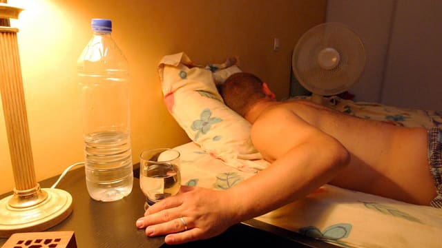 Mann liegt mit nacktem Oberkörper im Bett neben einem Ventilator Mann liegt mit nacktem Oberkörper im Bett neben einem Ventilator