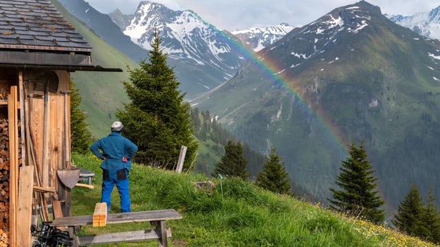 Eine Frau steht vor einer Alphütte und betrachtet einen Regenbogen. Eine Frau steht vor einer Alphütte und betrachtet einen Regenbogen.