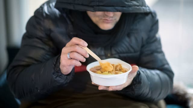 Ein Mann in einer dicken Kapuzenjacke löffelt eine Suppe aus.