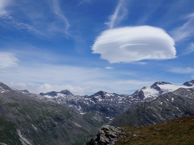 Blauer Himmel mit einer linsenförmigen Wolke mit mehreren Lagen. Darunter Gipfel des Hochgebirges mit etwa Schnee.