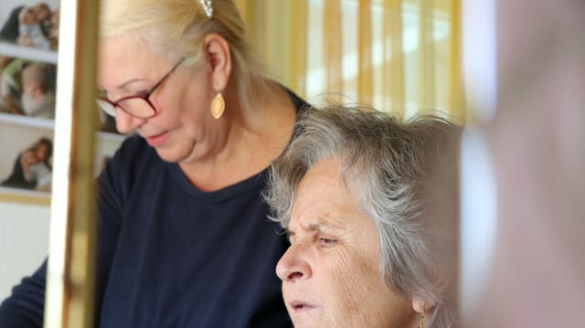 Im Vordergrund ist eine alte Frau zu sehen, also die Klientin, im Hintergrund die Pflegekraft aus Osteuropa Im Vordergrund ist eine alte Frau zu sehen, also die Klientin, im Hintergrund die Pflegekraft aus Osteuropa