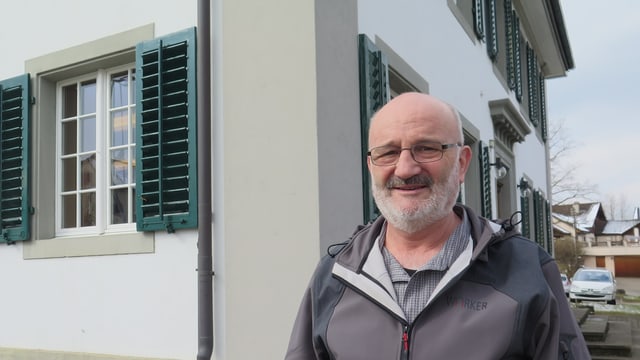 Ein Mann mit Brille, Glatze und weissem Bart steht vor einem Haus Ein Mann mit Brille, Glatze und weissem Bart steht vor einem Haus