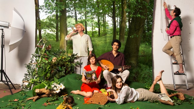 Die fünf Mitglieder der CapriConnection posieren als Gruppe vor einem Waldbild. Einer bläst ins Horn, einer spielt auf einer Laute, einer steht auf einer Leiter, eine liegt am Boden und eine sitzt und liest in einer Zeitschrift.