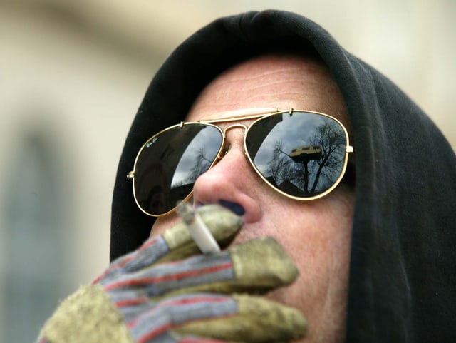 Flatz mit Kapuze und Sonnenbrille. Er trägt einen Arbeitshandschuh und zieht an einer Zigarette. In der Brille spiegelt sich die Installation «Belle Etage», ein Wohnwagen auf einem Baum. Flatz mit Kapuze und Sonnenbrille. Er trägt einen Arbeitshandschuh und zieht an einer Zigarette. In der Brille spiegelt sich die Installation «Belle Etage», ein Wohnwagen auf einem Baum.