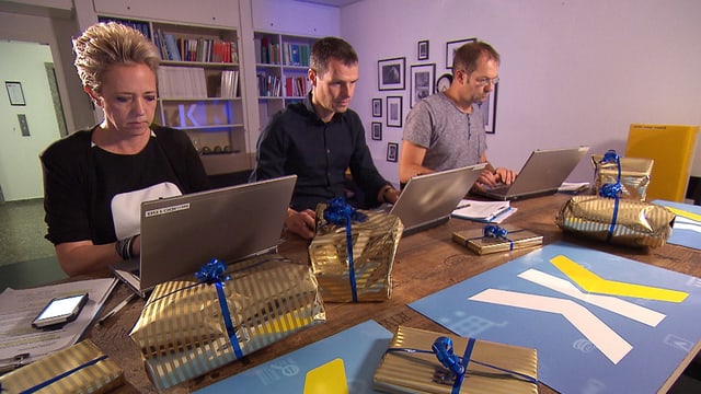 Zwei Männer und eine Frau sitzen je an einem Laptop, darum herum liegen Geschenke. Zwei Männer und eine Frau sitzen je an einem Laptop, darum herum liegen Geschenke.