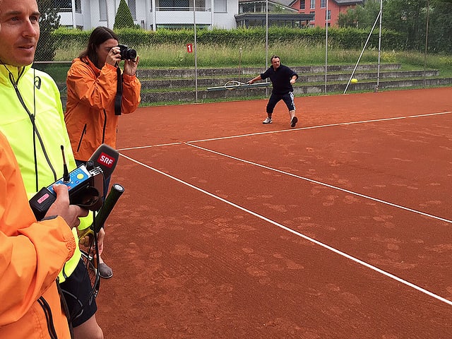 Viktor Röthlin steht mit Radioredaktoren neben dem Tennisplatz, Marco Rima spielt sich auf dem Platz ein.