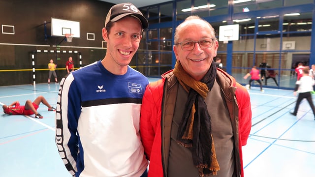 Zwei Männer in einer Turnhalle. Sie lächeln in die Kamera, im Hintergrund spielen Kinder Fussball. Zwei Männer in einer Turnhalle. Sie lächeln in die Kamera, im Hintergrund spielen Kinder Fussball.