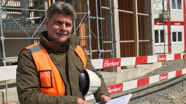 ... mit Warnweste und Helm unter dem Arm vor einem Gerüst und Baustellenabschrankungen. ... mit Warnweste und Helm unter dem Arm vor einem Gerüst und Baustellenabschrankungen.