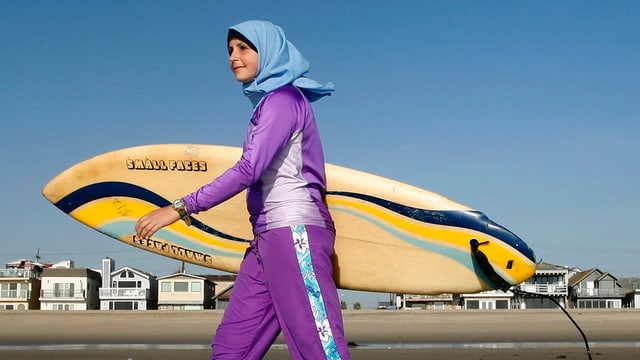 Eine Surferin in einem Burkini trägt ihr Brett an einem Strand. Eine Surferin in einem Burkini trägt ihr Brett an einem Strand.