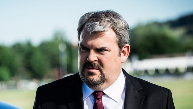 Mike Müller als Luc Conrad im dunklen Anzug mit roter Krawatte. Mike Müller als Luc Conrad im dunklen Anzug mit roter Krawatte.
