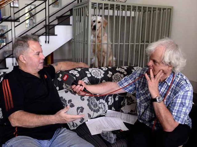 Zwei Männer sitzen auf einem Sofa. Einer hält dem anderen ein Mikrofon hin. Zwei Männer sitzen auf einem Sofa. Einer hält dem anderen ein Mikrofon hin.