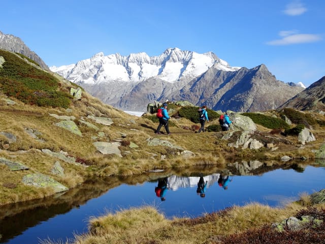 Berglandschaft und Wanderer bei Sonnenschein und blauem Himmel. Im Hintergrund verschneite Berge.
