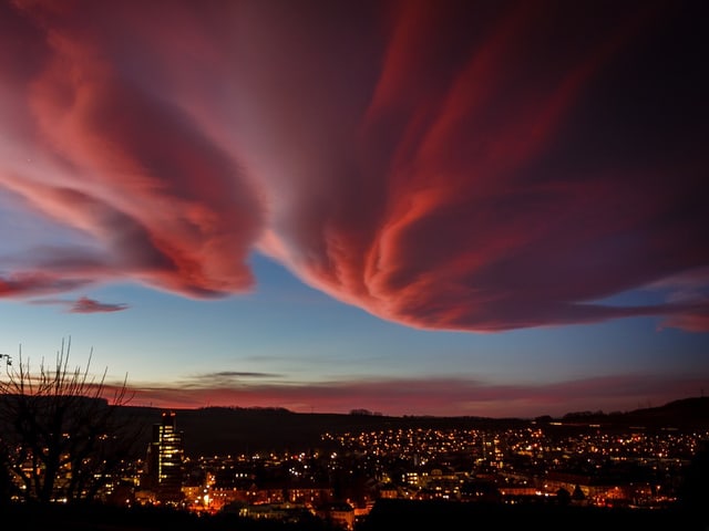 Riesige, rosarote Wolke am Himmel über einer Stadt. Es ist Abend und bereits dunkel. Die Wolke leuchtet genau so wie die Lichter der Stadt.