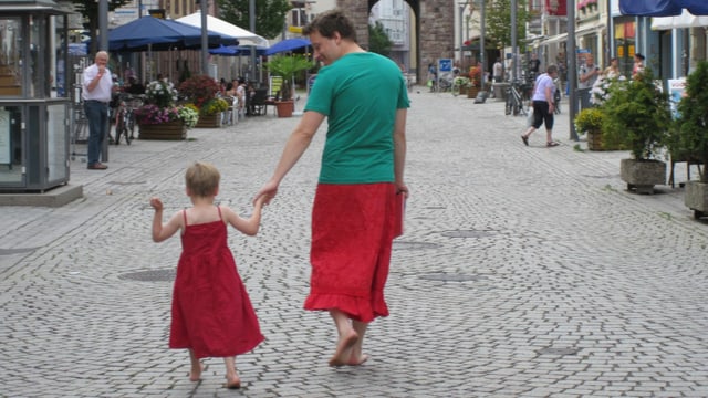 EIn Mann mit grünem Shirt und rotem Rock geht mit einem jungen in einem roten Kleid durch eine Strasse EIn Mann mit grünem Shirt und rotem Rock geht mit einem jungen in einem roten Kleid durch eine Strasse