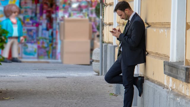 Mann im Anzug mit Smartphone und Kopfhörern lehnt an einer Hauswand. Mann im Anzug mit Smartphone und Kopfhörern lehnt an einer Hauswand.
