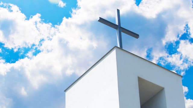 Ein moderner Kirchturm mit Kreuz vor blauem Himmel mit Wolken.