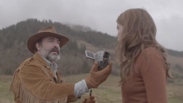 Ein Mann mit Hut und Lederjacke hält einer Frau eine Kamera entgegen. Ein Mann mit Hut und Lederjacke hält einer Frau eine Kamera entgegen.