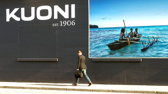 Werbeplakat der Firma mit polynesischen Ruderbootfahrern und einem Mann, der vor dem Plakat vorbeischreitet und zu Boden schaut. Werbeplakat der Firma mit polynesischen Ruderbootfahrern und einem Mann, der vor dem Plakat vorbeischreitet und zu Boden schaut.