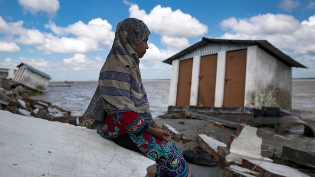 Ein Mädchen mit Kopftuch sitzt neben einem Haus, das zur Hälfte ins Meer gesunken ist. Ein Mädchen mit Kopftuch sitzt neben einem Haus, das zur Hälfte ins Meer gesunken ist.
