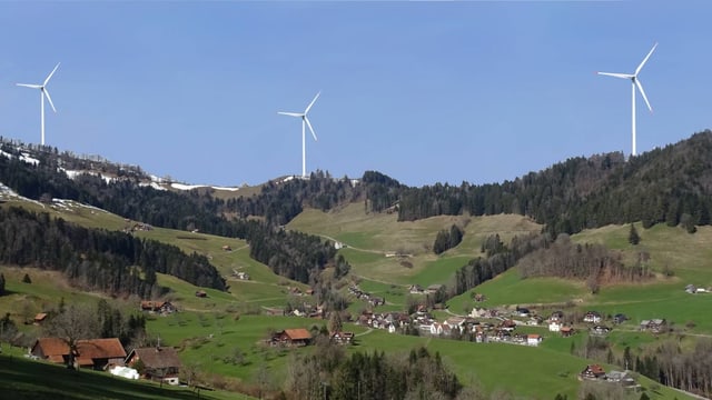 Drei Windräder stehen auf einer Bergkrete, im Vordergrund ein dorf in grüner Landschaft Drei Windräder stehen auf einer Bergkrete, im Vordergrund ein dorf in grüner Landschaft