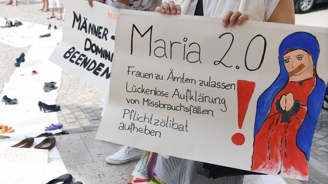 Eine Frau hält ein Protestschild, auf dem eine Maria mit zugeklebtem Mund zu sehen ist. Eine Frau hält ein Protestschild, auf dem eine Maria mit zugeklebtem Mund zu sehen ist.