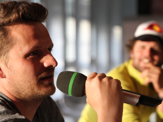 Jon Cavegn spricht in ein Mikrofon.  Ein Diskussionsteilnehmer hört im Hintergrund zu.