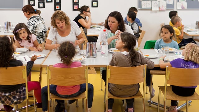 In einer Kinderkrippe sitzen mehrere Kinder und zwei Betreuerinnen an einem Esstisch In einer Kinderkrippe sitzen mehrere Kinder und zwei Betreuerinnen an einem Esstisch