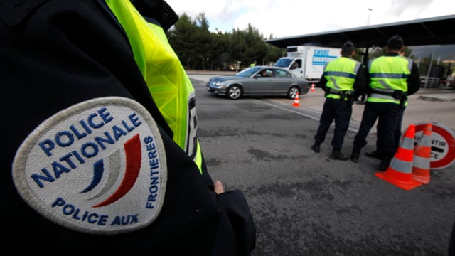 Der Arm eines Polizisten an einer französischen Grenze, im Hintergrund weitere Polizisten und Fahrzeuge.
