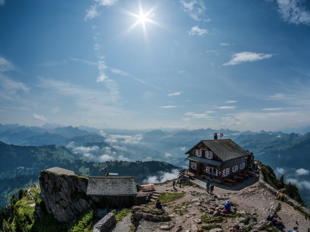 Berggipfel mit Hütte und ein paar Wanderern. Der Himmel ist hellblau und die Sonne scheint.