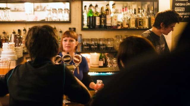 Leute sitzen an einer Bar, eine Kellnerin zapft ein Bier.