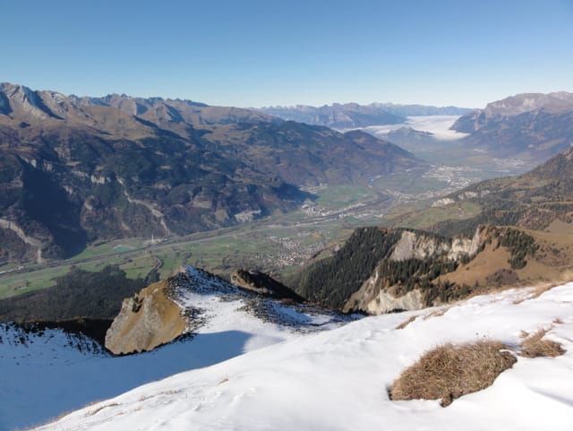 Auf dem Berggipfel liegt zum Teil noch Schnee. Unten im Tal schlängelt sich der Rhein talabwärts und taucht im St. Gallerrheintal in den Hochnebel ein. Darüber ein wolkenloser Himmel.