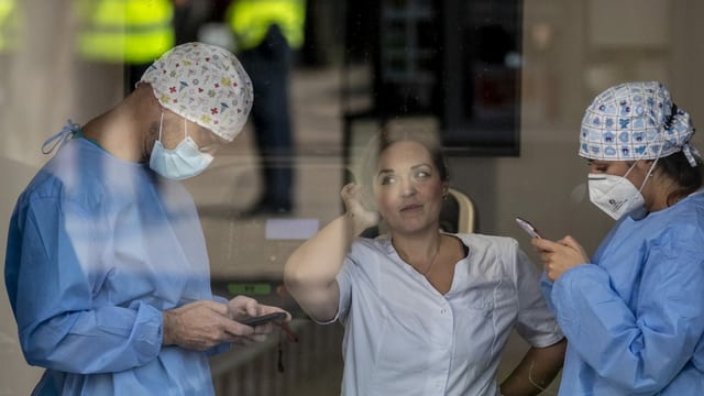 Zwei Klinikmitarbeiter tragen Masken und sind am Handy, eine Pflegeangestellte ohne Maske lächelt sie an.