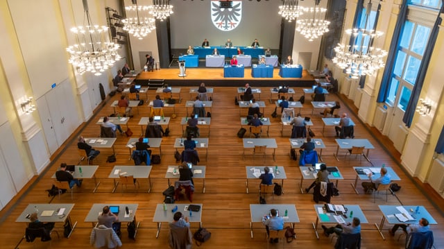 Ratssitzung in einem grossen Saal, alle Personen an eigenem Tisch. Ratssitzung in einem grossen Saal, alle Personen an eigenem Tisch.