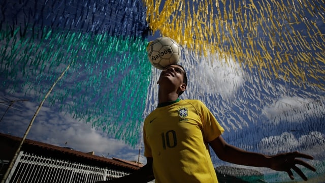 Symbolbild: Junger Mann mit Brasilien-Shirt balanciert einen Fussball auf dem Kopf.