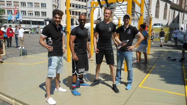 Vier Sportler in schwarzen T-Shirts posieren vor einem gelben Klettergerüst. Vier Sportler in schwarzen T-Shirts posieren vor einem gelben Klettergerüst.