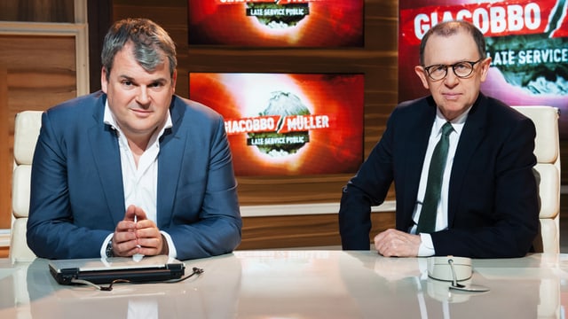 Mike Müller in weissem Hemd und blauem Blazer und Viktor Giacobbo in weissem Hemd, Kravatte und dunklem Blazer sitzen an einem Blanken Tisch. Sie blicken den Betrachter an. Im Hintergrund zu sehen: Das Logo der TV-Sendung «Giacobbo / Müller». Mike Müller in weissem Hemd und blauem Blazer und Viktor Giacobbo in weissem Hemd, Kravatte und dunklem Blazer sitzen an einem Blanken Tisch. Sie blicken den Betrachter an. Im Hintergrund zu sehen: Das Logo der TV-Sendung «Giacobbo / Müller».