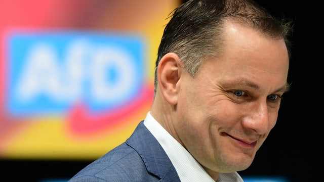 Tino Chrupalla lächelnd vor einem AfD-Logo. Tino Chrupalla lächelnd vor einem AfD-Logo.