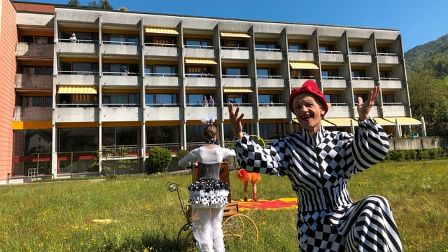 Ein Clown kniet in einer Wiese, im Hintergrund ist ein Altersheim, die Leute stehen auf den Balkonen. Ein Clown kniet in einer Wiese, im Hintergrund ist ein Altersheim, die Leute stehen auf den Balkonen.