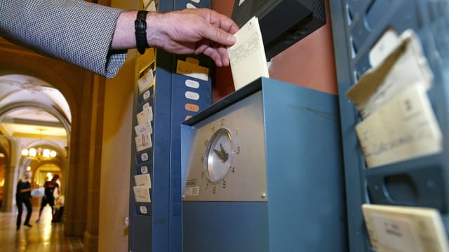 Eine Hand führt eine Zeiterfassungskarte in eine Stempleuhr ein. Eine Hand führt eine Zeiterfassungskarte in eine Stempleuhr ein.