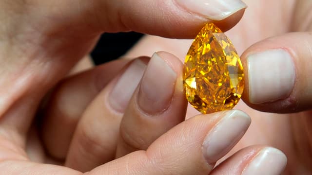 Der orange farbene Diamant zwischen Damenfingern. Seine Form jene eines Tropfens. Der orange farbene Diamant zwischen Damenfingern. Seine Form jene eines Tropfens.