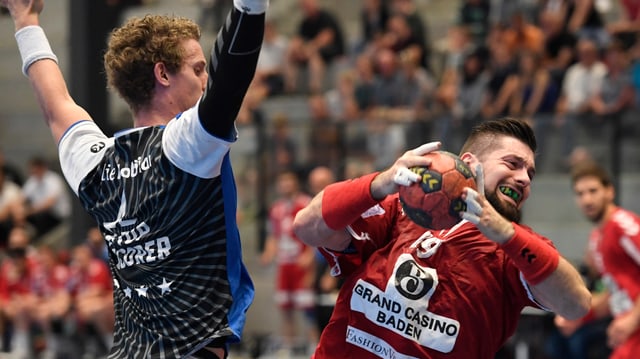 Mann in rotem Shirt hält Handball, Mann im schwarzen Shirt reisst Arme in die Luft. Mann in rotem Shirt hält Handball, Mann im schwarzen Shirt reisst Arme in die Luft.
