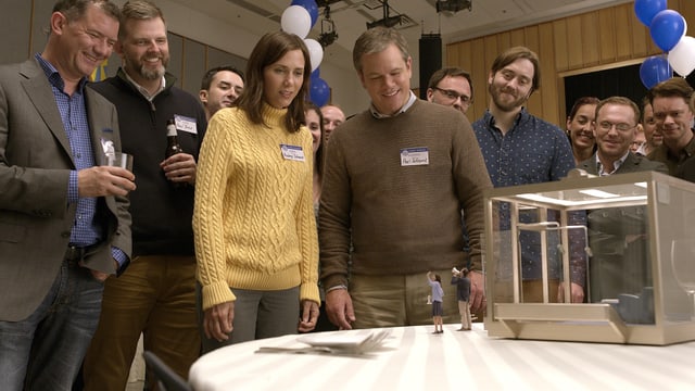 Eine Gruppe normalgrosser Menschen steht um einen Tisch. Sie alle tragen Namensschilder, im Hintergrund sind Ballons zu sehen. Auf dem Tisch stehen ein winziger Mann und eine winzige Frau, die durch ein Megaphon zu den Grossen emporrufen. Eine Gruppe normalgrosser Menschen steht um einen Tisch. Sie alle tragen Namensschilder, im Hintergrund sind Ballons zu sehen. Auf dem Tisch stehen ein winziger Mann und eine winzige Frau, die durch ein Megaphon zu den Grossen emporrufen.