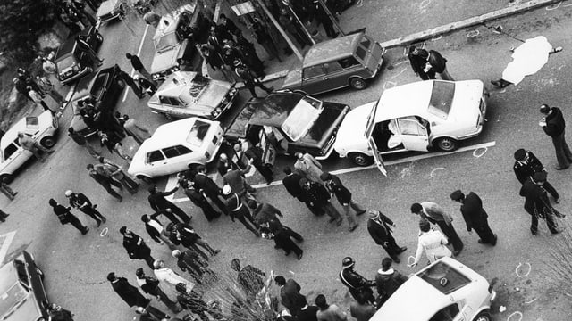 Mehrere Leute stehen um ein paar Autors herum, die quer auf einer Strasse stehen. Mehrere Leute stehen um ein paar Autors herum, die quer auf einer Strasse stehen.