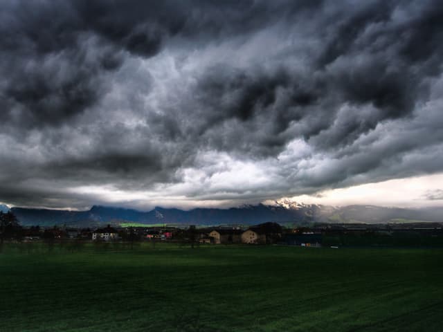 Dunke bis hellgraue Wolken. Darunter Häuser und Wiesen.