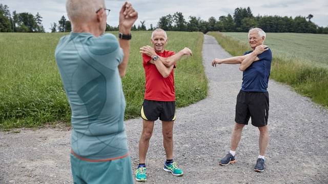 Drei ältere Luzerner Männer in Jogging-Anzügen stretchen ihre Arme. Drei ältere Luzerner Männer in Jogging-Anzügen stretchen ihre Arme.