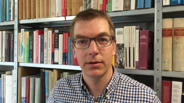 Mann mit Brille und kariertem Hemd vor einem Gestell mit ganz vielen Büchern Mann mit Brille und kariertem Hemd vor einem Gestell mit ganz vielen Büchern