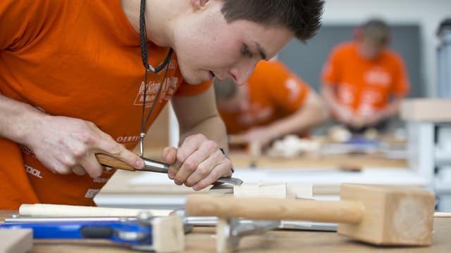 Lehrling bearbeitet an seinem Arbeitsplatz mit einem Werkzeug ein Stück Holz. Lehrling bearbeitet an seinem Arbeitsplatz mit einem Werkzeug ein Stück Holz.