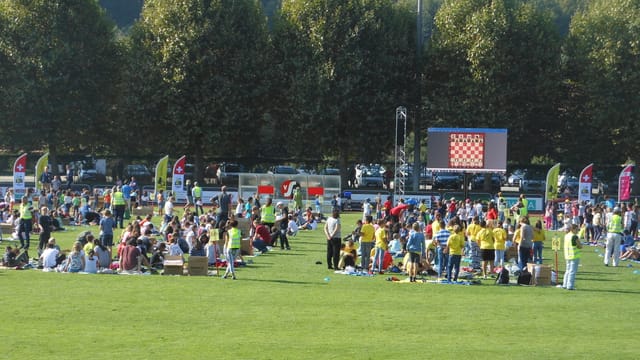 Man sieht viele Schüler und Schülerinnen, die auf dem Sportplatz stehen und sitzen und auf die Grossleinwand schauen Man sieht viele Schüler und Schülerinnen, die auf dem Sportplatz stehen und sitzen und auf die Grossleinwand schauen