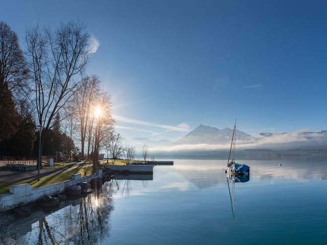 Die Sonne und die Alpen spiegeln sich auf der glatten Seeoberfläche. Darüber blauer Himmel.